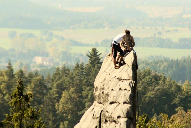 two people on top of a rock face jutting out from mountain overlooking a forest