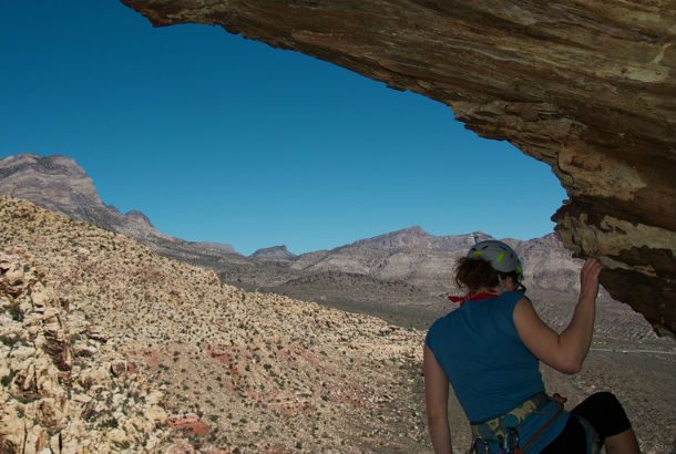 woman hangs from fingertips from the side of a mountain under an arch